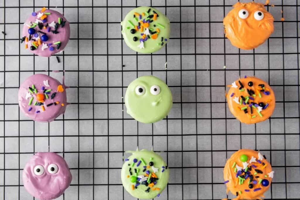 Nine colorful, round cookies decorated with candy eyes and sprinkles arranged in a grid pattern on a black cooling rack.