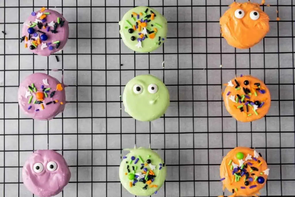 Nine colorful, round cookies decorated with candy eyes and sprinkles arranged in a grid pattern on a black cooling rack.