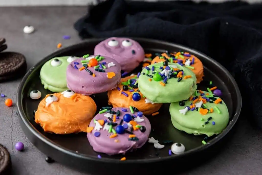 A black plate with Oreo cookies covered in green, orange, and purple icing, decorated with sprinkles and candy eyes, sits on a gray surface.
