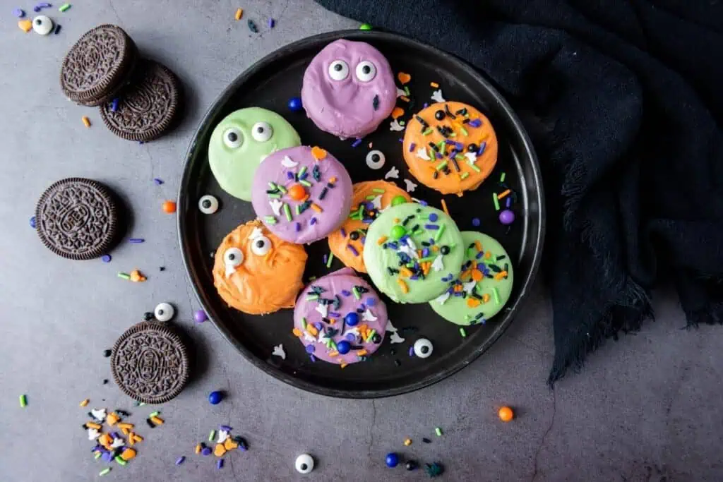 A black plate holds colorful, coated cookies with candy eyes and sprinkles; Oreo cookies and extra sprinkles are scattered on a gray surface.