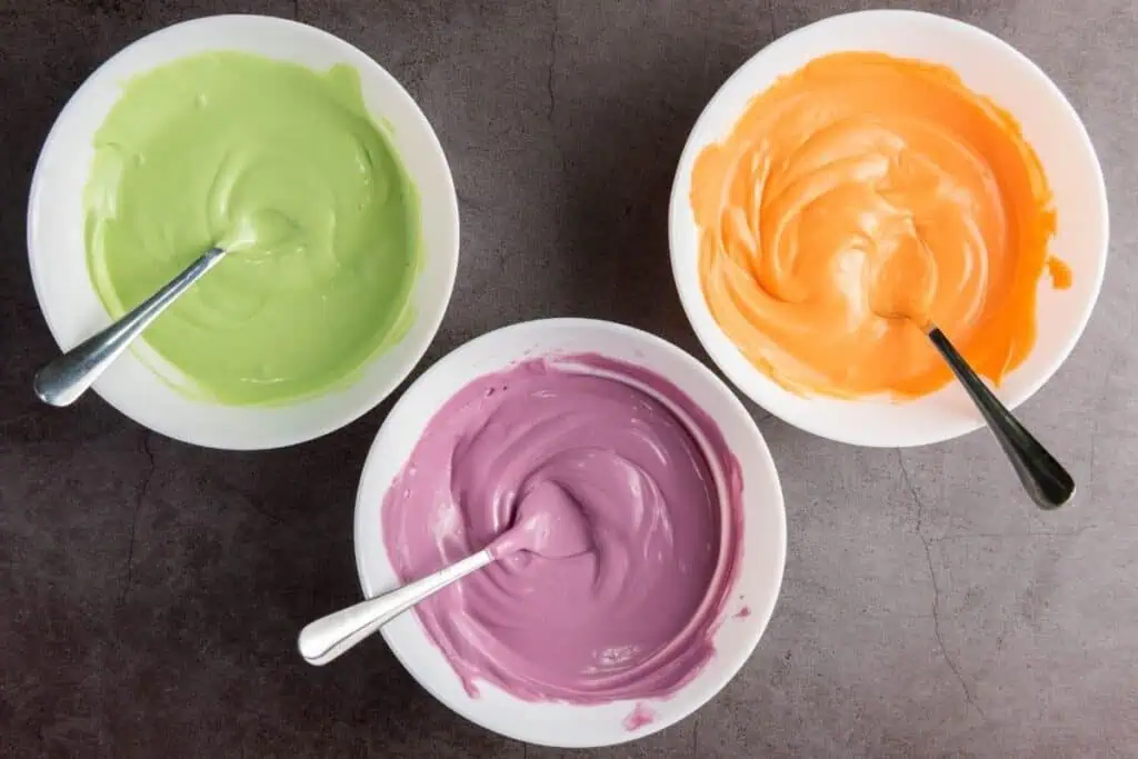Three bowls filled with colorful melted candies: green, orange, and purple, each with a spoon, placed on a dark countertop.