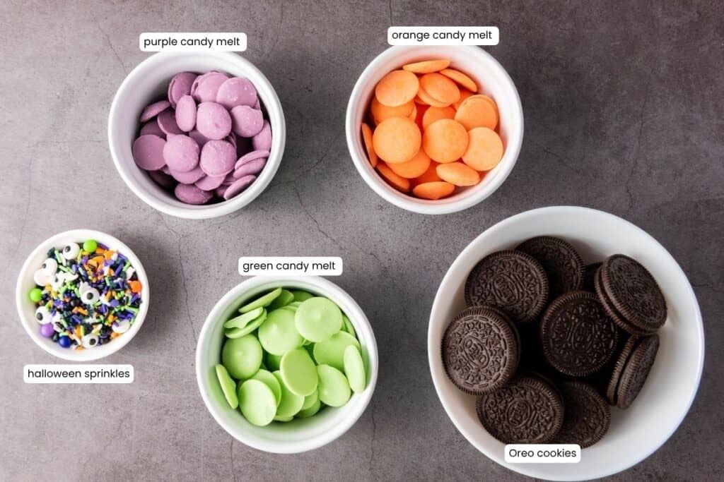 Five bowls containing purple candy melts, orange candy melts, green candy melts, Halloween sprinkles, and Oreo cookies, all labeled on a gray surface.