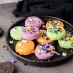 A black plate with Halloween-themed cookies coated in orange, purple, and green, decorated with sprinkles and candy eyes. Some Oreo cookies and sprinkles are scattered nearby.