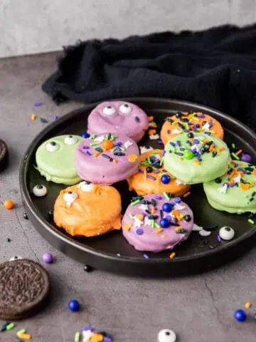 A black plate with Halloween-themed cookies coated in orange, purple, and green, decorated with sprinkles and candy eyes. Some Oreo cookies and sprinkles are scattered nearby.