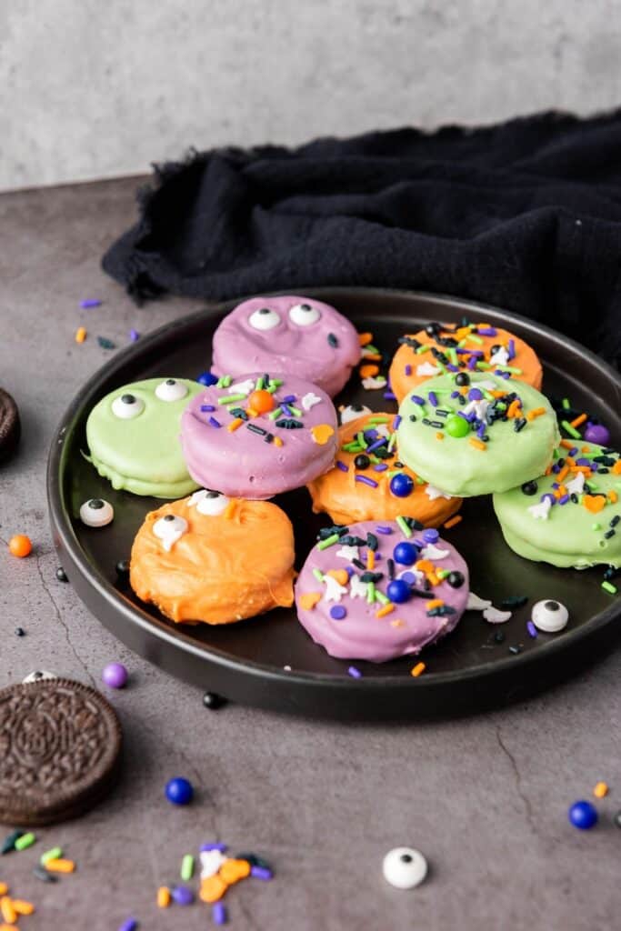 A black plate with Halloween-themed cookies coated in orange, purple, and green, decorated with sprinkles and candy eyes. Some Oreo cookies and sprinkles are scattered nearby.