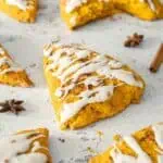 Pumpkin Spice Scones with Vanilla Glaze are arranged on a light surface, with some whole and one halved, surrounded by cinnamon sticks and star anise.