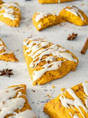 Pumpkin Spice Scones with Vanilla Glaze are arranged on a light surface, with some whole and one halved, surrounded by cinnamon sticks and star anise.