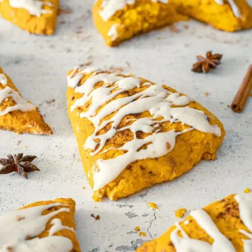 Pumpkin Spice Scones with Vanilla Glaze are arranged on a light surface, with some whole and one halved, surrounded by cinnamon sticks and star anise.