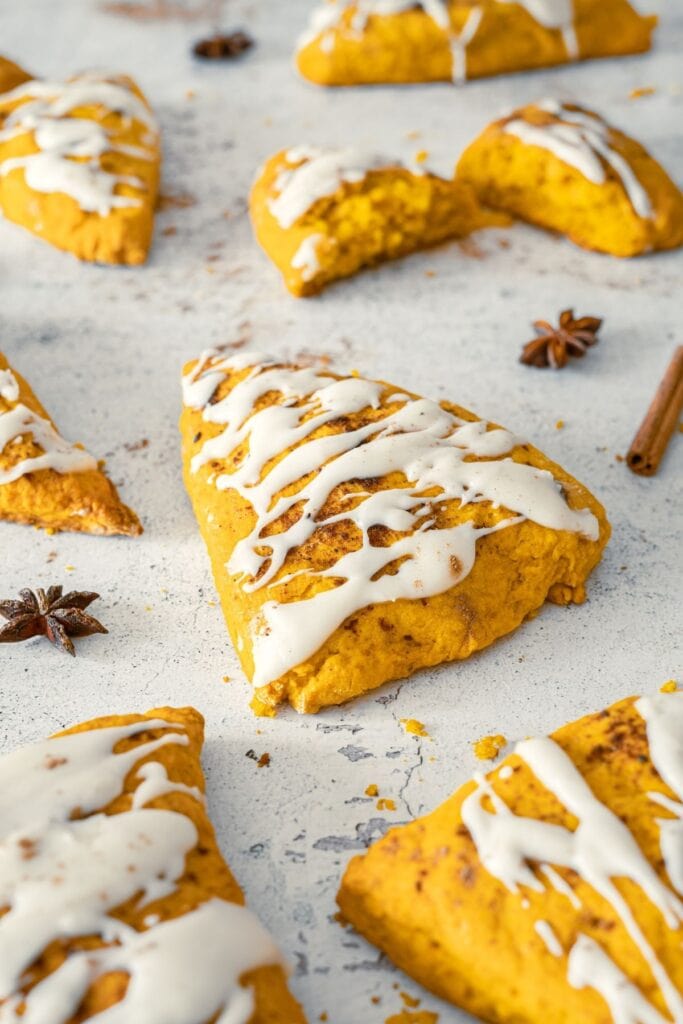 Pumpkin Spice Scones with Vanilla Glaze are arranged on a light surface, with some whole and one halved, surrounded by cinnamon sticks and star anise.