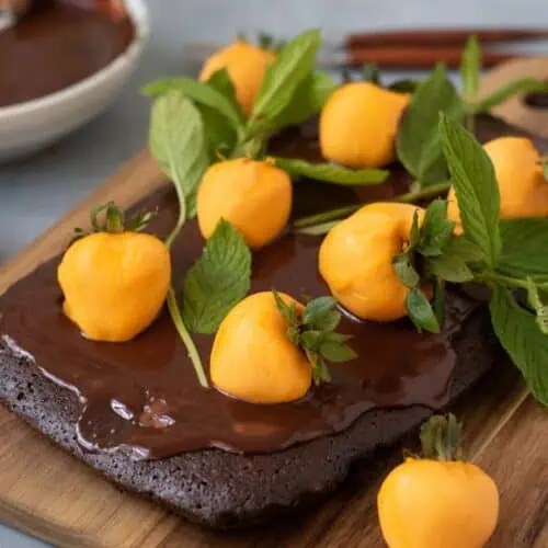 A chocolate cake topped with glossy chocolate icing, yellow strawberries, and fresh green mint leaves on a wooden board.