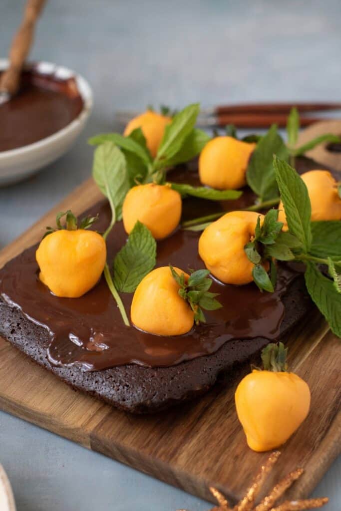 A chocolate cake topped with glossy chocolate icing, yellow strawberries, and fresh green mint leaves on a wooden board.