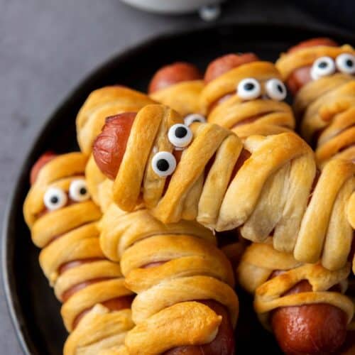 Several hot dogs wrapped in crescent dough to look like mummies, with candy eyes added, are arranged on a black plate. A small bowl of dipping sauce is in the background.