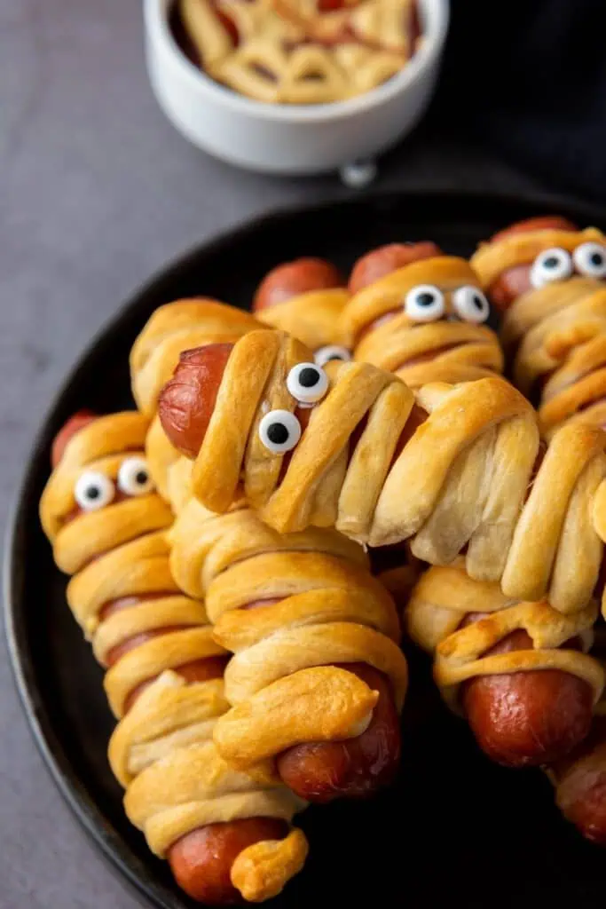 Several hot dogs wrapped in crescent dough to look like mummies, with candy eyes added, are arranged on a black plate. A small bowl of dipping sauce is in the background.