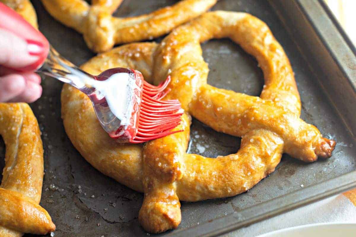 A hand uses a red silicone brush to apply butter or glaze to a baked soft pretzel on a baking sheet.