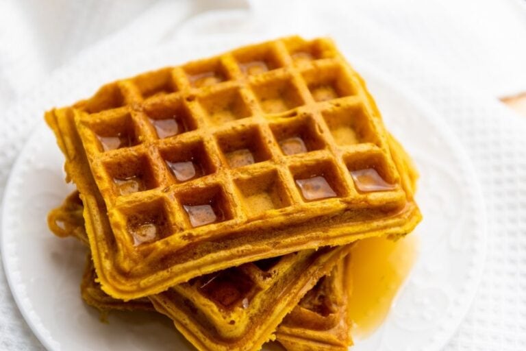 Three square waffles stacked on a white plate, with syrup pooled in some of the squares and dripping onto the plate.