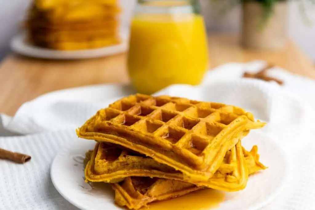 A plate with three stacked waffles drizzled with syrup, with a glass of orange juice and more waffles in the background.