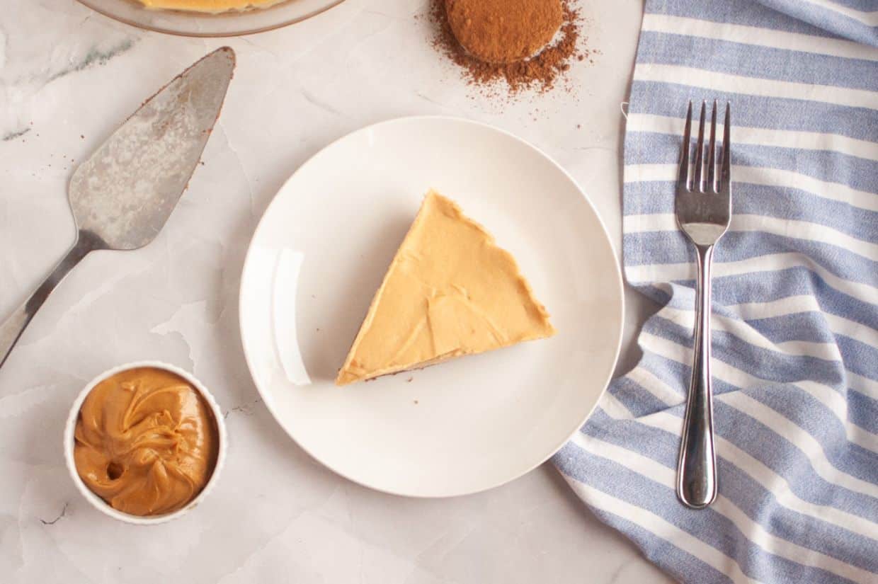 A slice of Peanut Butter & Chocolate Pie on a white plate, with a fork, a small bowl of peanut butter, a baking tool, and a striped napkin nearby.