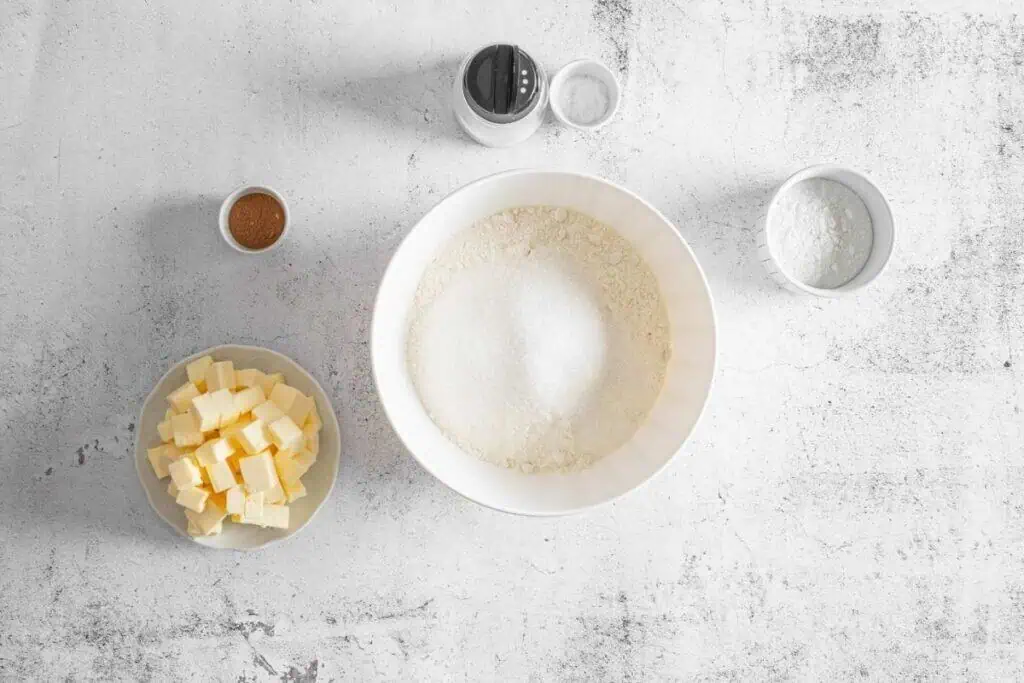 Baking ingredients on a white surface, including a bowl with flour and sugar, a dish of cubed butter, and small bowls of pumpkin pie spice, baking powder, and salt.