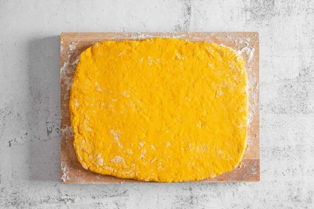 A rectangular sheet of orange dough is rolled out on a floured wooden board, placed on a light-colored textured surface.