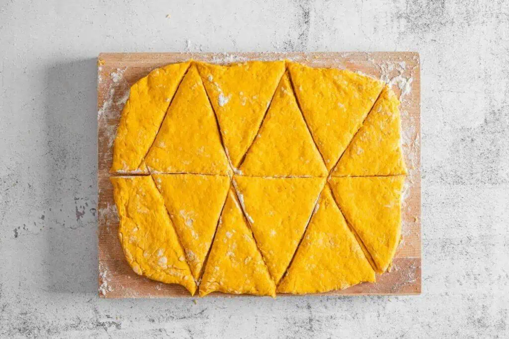 Rectangular sheet of orange dough cut into triangle pieces on a floured wooden board, viewed from above.