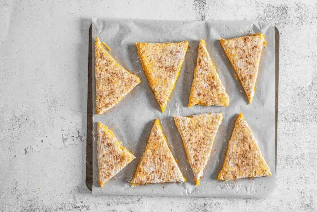 Eight triangular pieces of uncooked pastry, dusted with pumpkin pie spice, arranged on a sheet of parchment paper atop a baking tray.