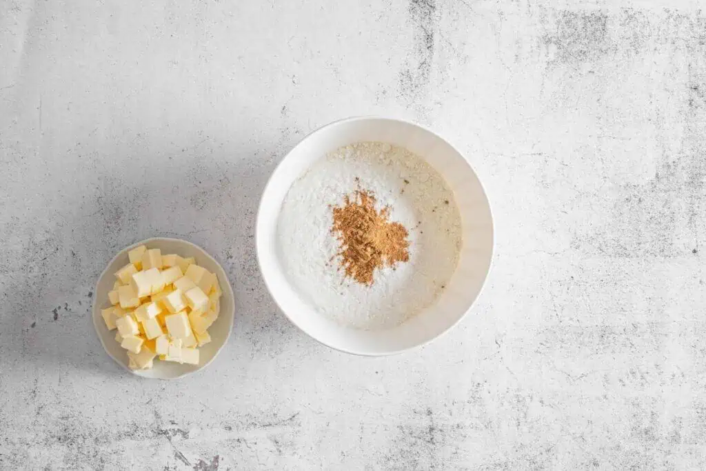 A bowl of cubed butter beside a large bowl containing all the dry ingredients on a light textured surface.