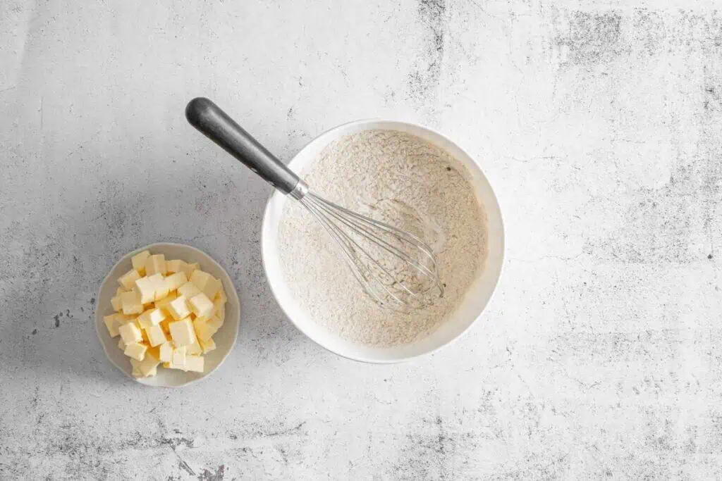A bowl of dry ingredients mix with a whisk inside next to a small bowl of cubed butter on a light textured surface.
