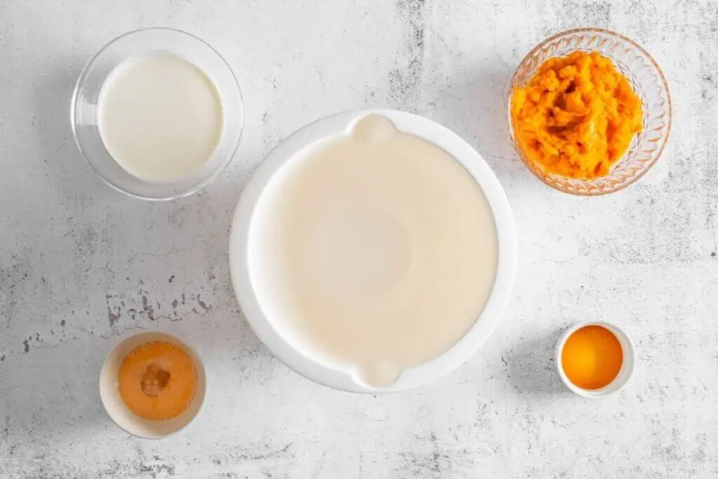 Four bowls containing cream, pureed pumpkin, egg, and vanilla are arranged alongside an empty mixing bowl on a light, textured surface.