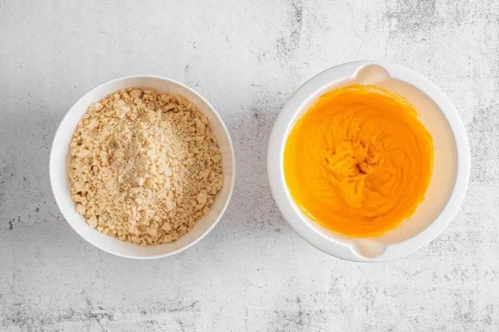 Two white bowls on a light surface: one filled with crumbled dry mixture, the other containing smooth orange batter.