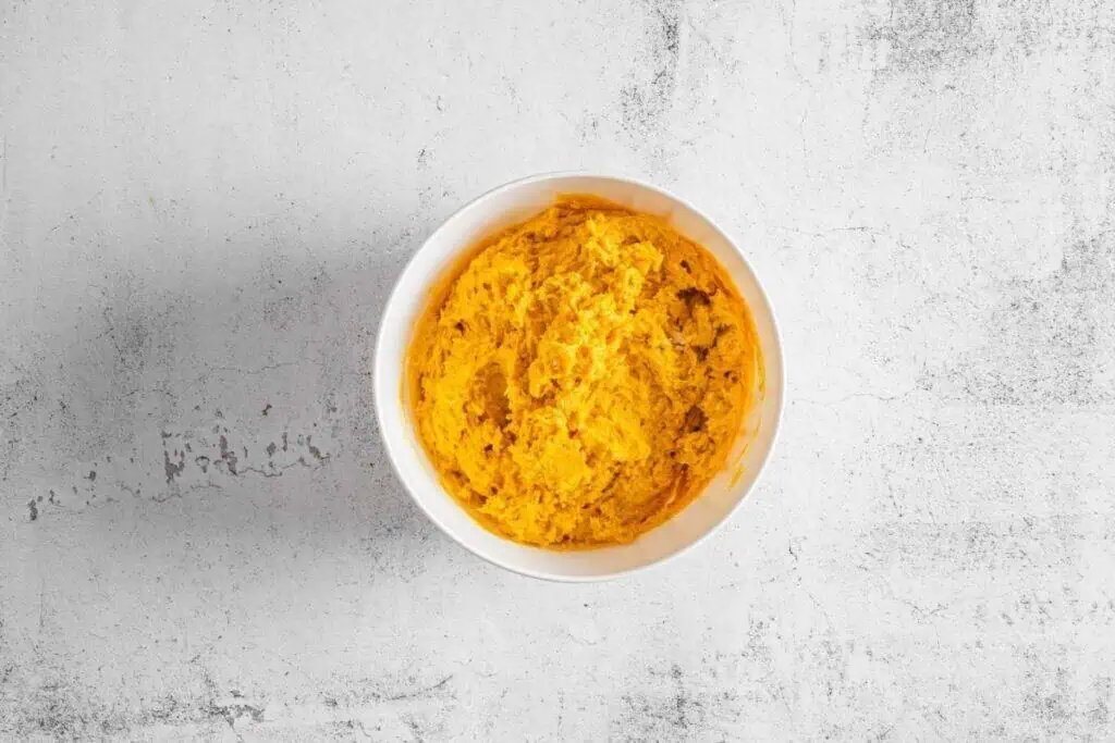 A white bowl filled with an orange dough is placed on a light textured surface.