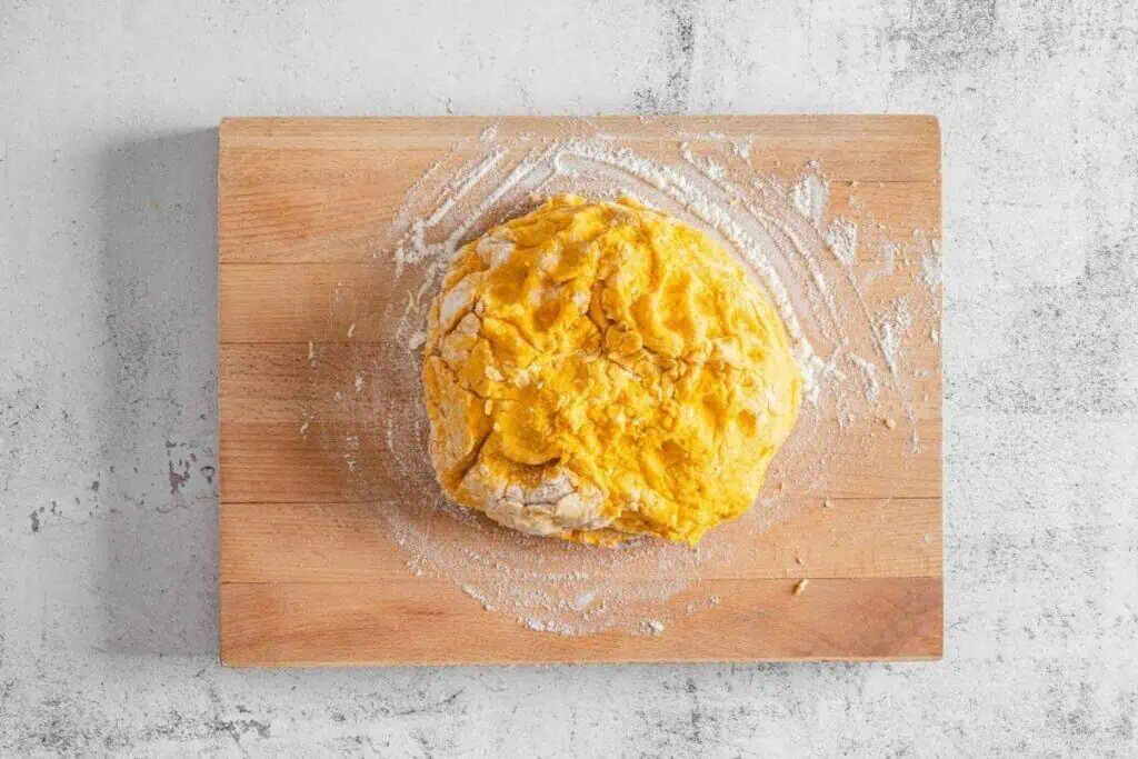 A ball of yellow dough sits on a floured wooden cutting board atop a light gray textured surface.