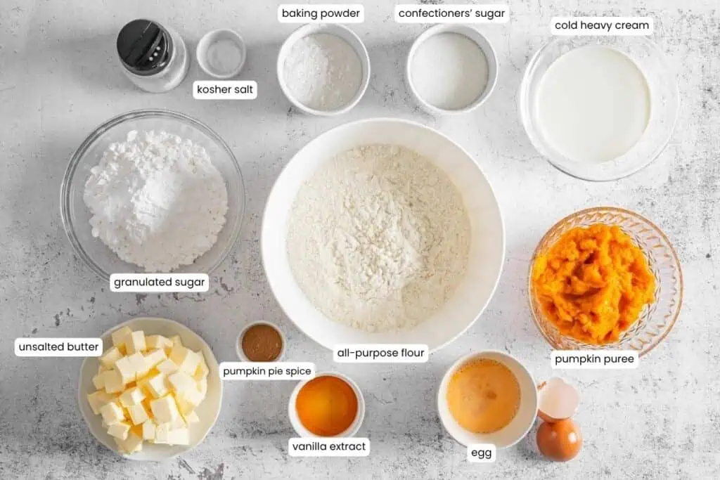 Various baking ingredients, including flour, sugars, pumpkin puree, butter, egg, cream, vanilla extract, spices, baking powder, and salt, arranged in separate bowls on a white surface.