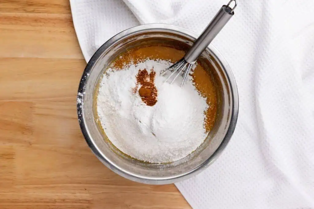 A metal bowl with flour, ground cinnamon, salt, and liquid ingredients being mixed with a whisk on a wooden surface next to a white cloth.