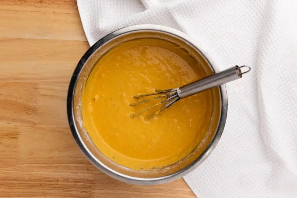 A metal bowl filled with orange batter and a whisk sits on a wooden surface next to a white textured cloth.