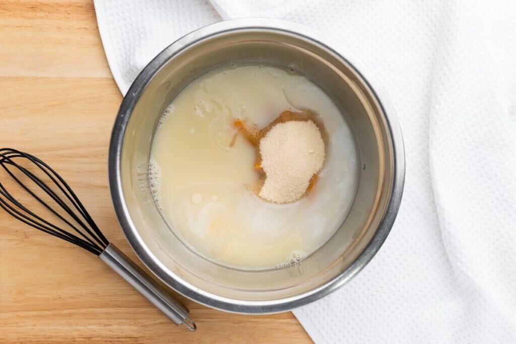 A metal mixing bowl with milk, pumpkin puree, melted butter, and sugar, next to a black wire whisk on a wooden surface with a white towel.