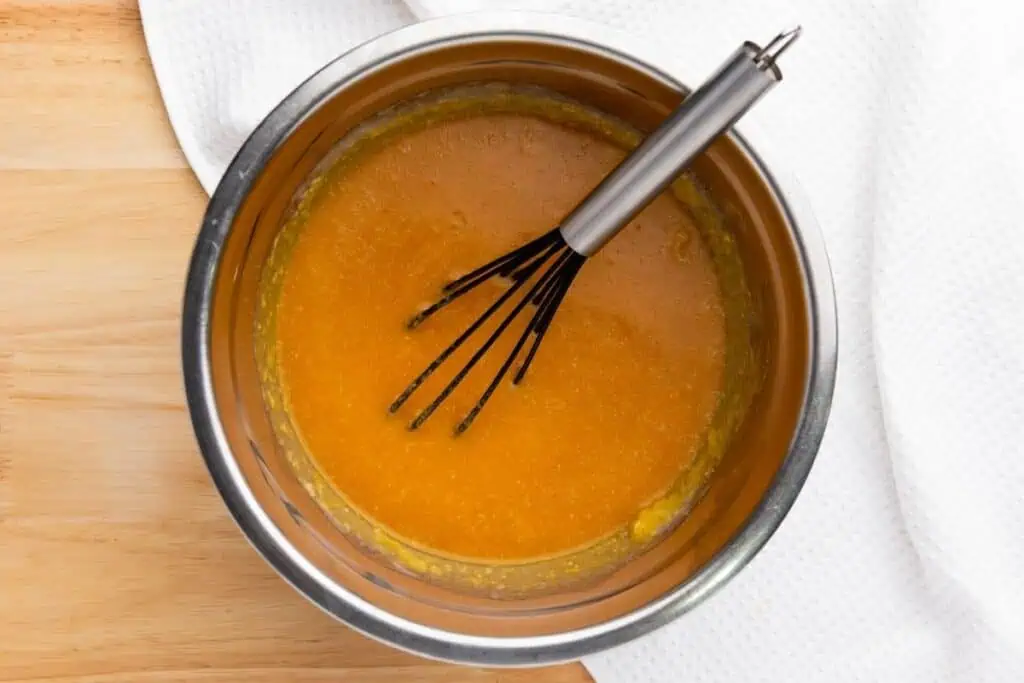 A metal bowl with orange batter being mixed with a whisk, placed on a wooden surface next to a white textured cloth.
