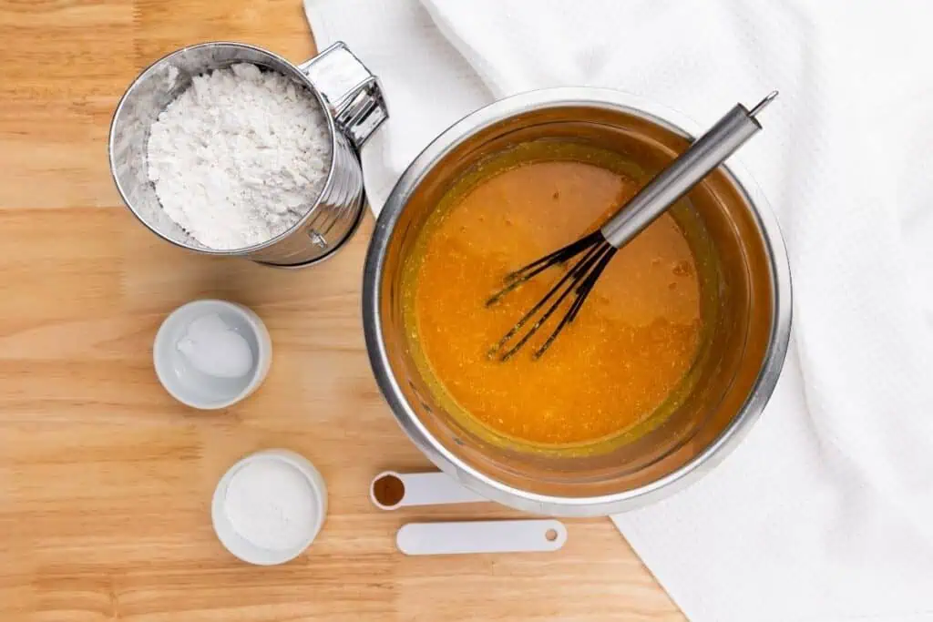 Metal bowl with orange batter and a whisk, flour, small bowls of baking powder and soda, and measuring spoons with cinnamon and salt on a wooden surface next to a white cloth.