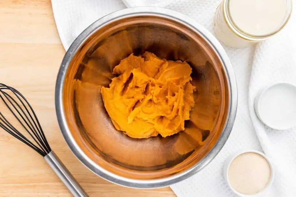 A metal mixing bowl containing pumpkin puree sits on a wooden surface next to a whisk, a jar, a small bowl, and a white towel.