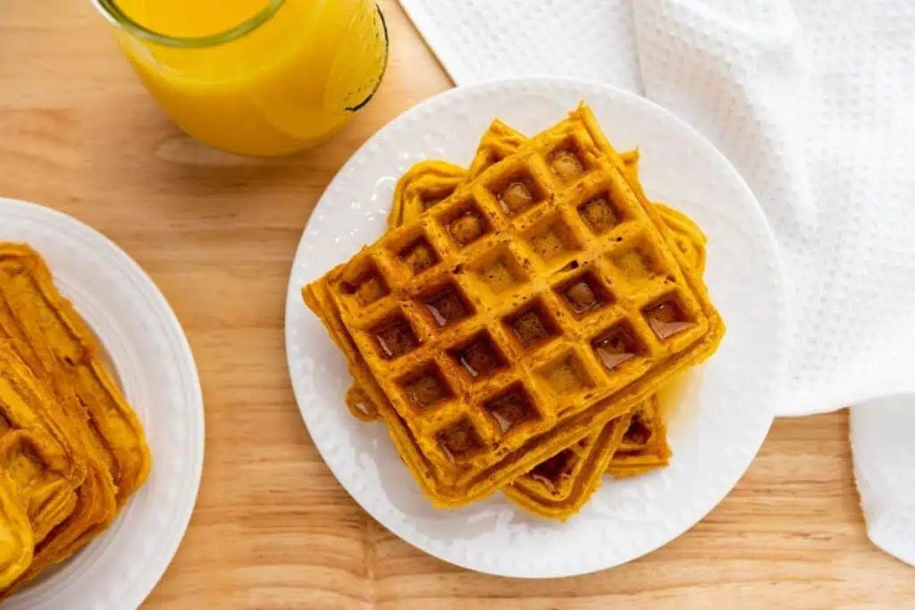A plate of square waffles with syrup sits on a wooden table next to a glass of orange juice and a white textured napkin.