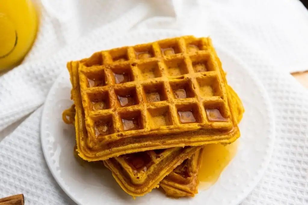 A stack of square waffles with syrup on a white plate, placed on a textured white cloth.