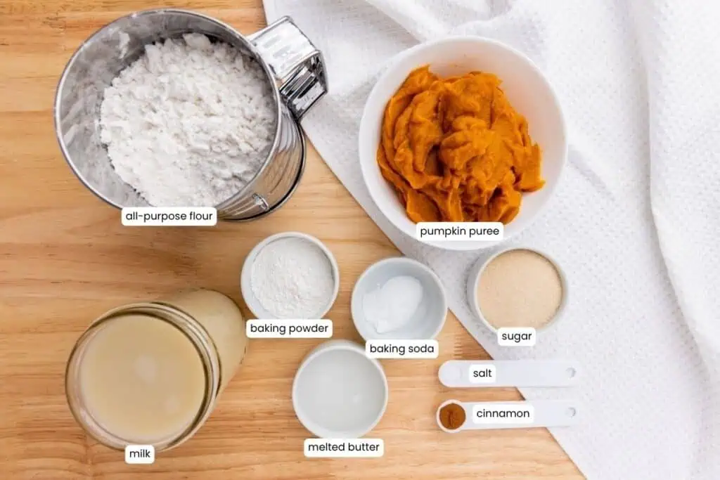 A top-down view of labeled baking ingredients in bowls and measuring spoons, including flour, pumpkin puree, milk, baking powder, baking soda, butter, sugar, salt, and cinnamon.