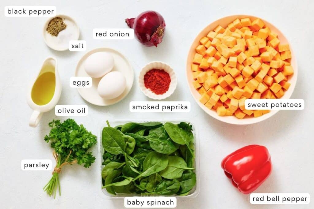 A variety of labeled ingredients on a white surface, including sweet potatoes, eggs, red onion, red bell pepper, baby spinach, parsley, olive oil, salt, black pepper, and smoked paprika.