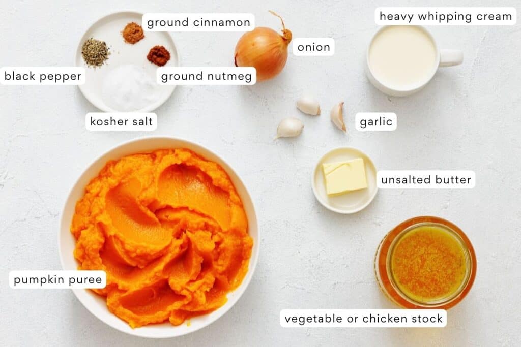 Ingredients for pumpkin soup are arranged on a white surface, including pumpkin puree, spices, onion, garlic, butter, cream, and broth.