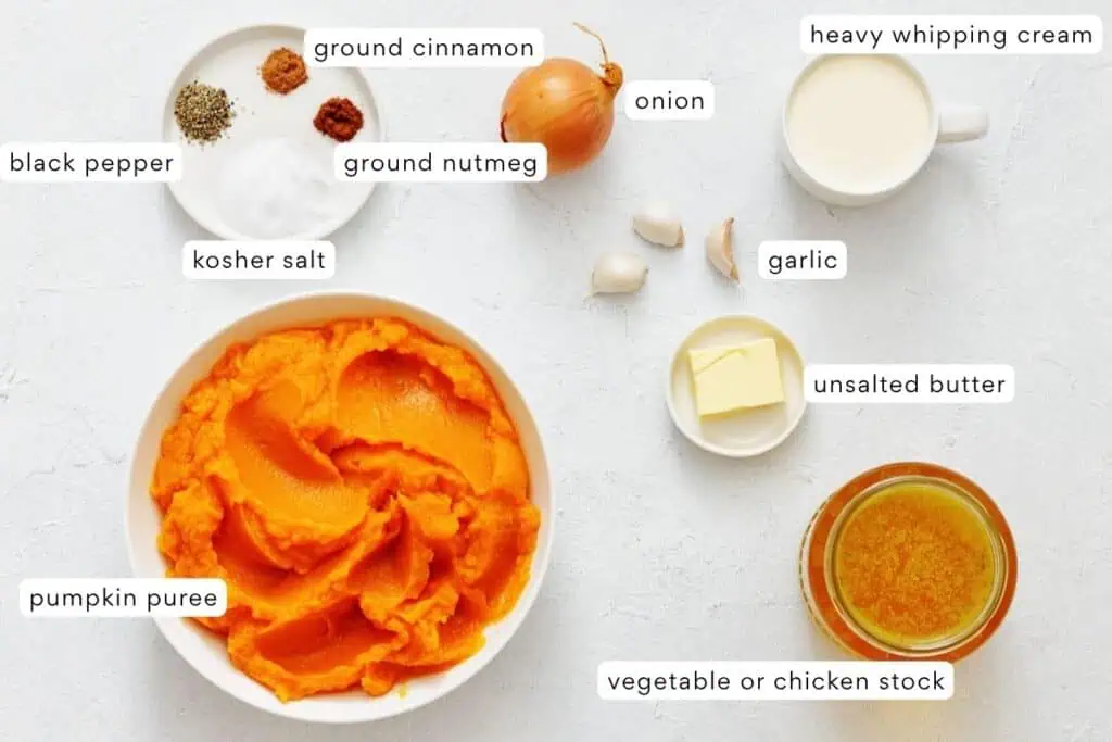 Ingredients for pumpkin soup are arranged on a white surface, including pumpkin puree, spices, onion, garlic, butter, cream, and broth.