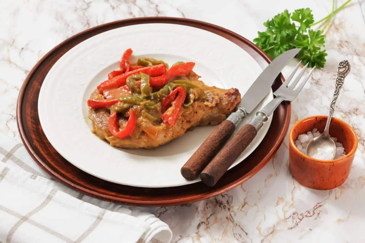 Smothered Pork Chops on a white plate, served with a fork and knife, next to a small bowl of salt and a sprig of parsley.