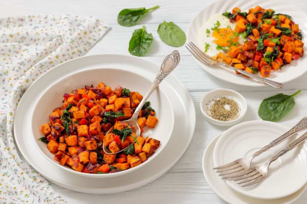 A bowl of sweet potato hash with spinach and red onions, served with a side plate, fork, and a dish of ground pepper on a white table.