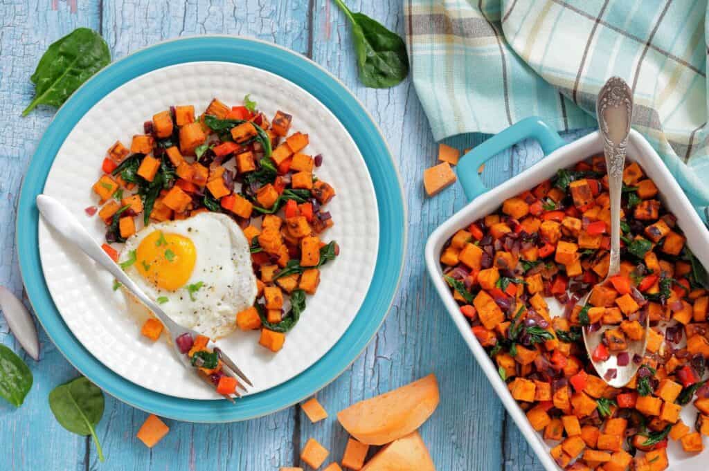 A plate with a fried egg and a serving of sweet potato hash next to a baking dish filled with more hash on a blue wooden table with a cloth napkin.