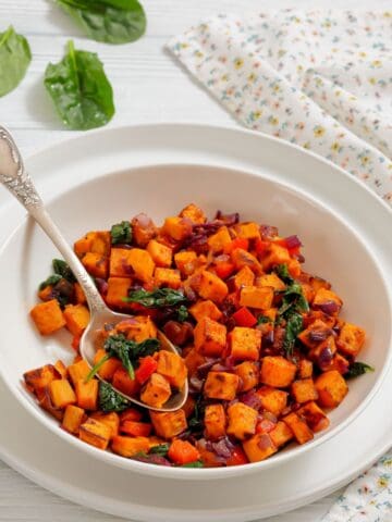 A white bowl filled with a sweet potato hash containing diced sweet potatoes, red onions, spinach, and red bell peppers, with a spoon resting inside.
