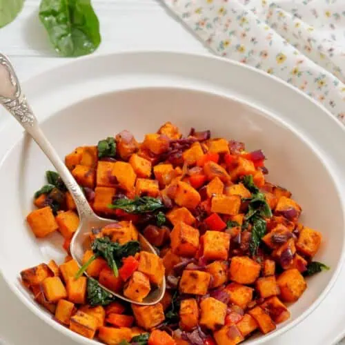 A white bowl filled with a sweet potato hash containing diced sweet potatoes, red onions, spinach, and red bell peppers, with a spoon resting inside.