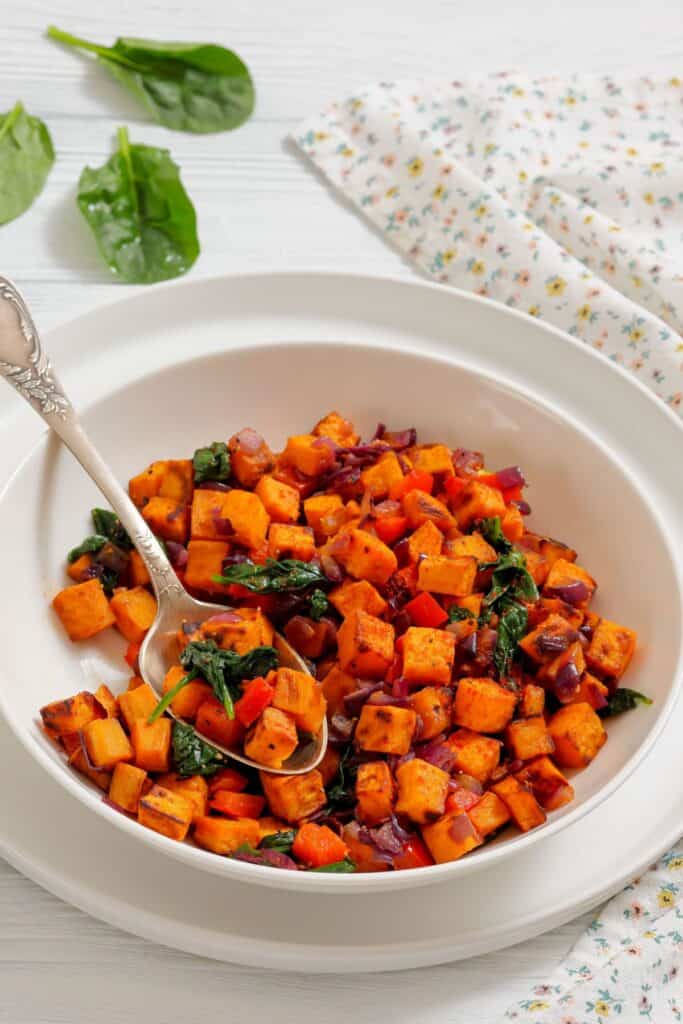 A white bowl filled with a sweet potato hash containing diced sweet potatoes, red onions, spinach, and red bell peppers, with a spoon resting inside.